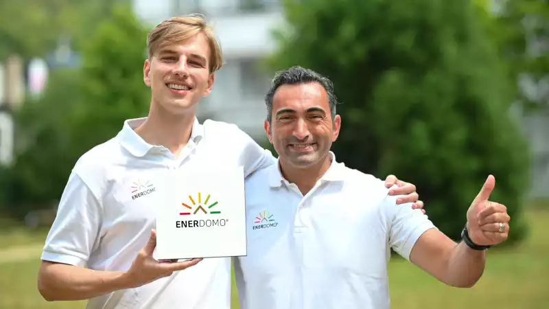 Zwei Männer in weißen Poloshirts mit ENERDOMO-Logo lächeln im Grünen und halten ein Firmenschild.